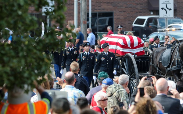A military funeral procession moves through a crowded street, featuring a flag-draped casket carried on a horse-drawn caisson. Uniformed soldiers in dress uniforms escort the procession on foot while mounted soldiers ride alongside on dark horses. A large crowd of civilians lines the street to pay their respects, with safety personnel in high-visibility vests managing the crowd.