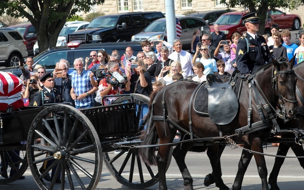A military funeral procession is taking place on a public street, featuring a flag-draped casket carried on a horse-drawn caisson pulled by six dark horses. Uniformed military personnel walk alongside the caisson as an honor guard, while a large crowd of civilians lines the street to pay their respects. The scene reflects a formal military burial ceremony consistent with U.S. Army funeral honors.