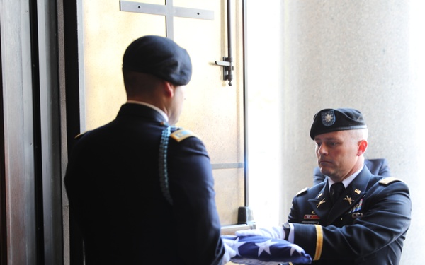 A military funeral ceremony is taking place, with two U.S. Army soldiers in dress uniforms performing a flag folding or presentation ritual. A casket is visible in the lower left corner, and civilian attendees are visible in the background. The setting appears to be a formal memorial building or chapel entrance with stone columns.