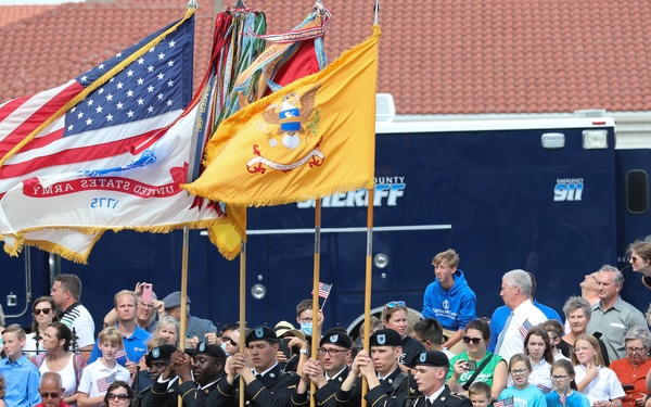 A U.S. Army color guard presents flags during what appears to be a patriotic parade or ceremony, likely Memorial Day or Independence Day. Soldiers in dress uniforms carry the American flag, the United States Army flag (dated 1775), and additional military colors. A large crowd of civilians, including families and children, lines the street to observe the event.
