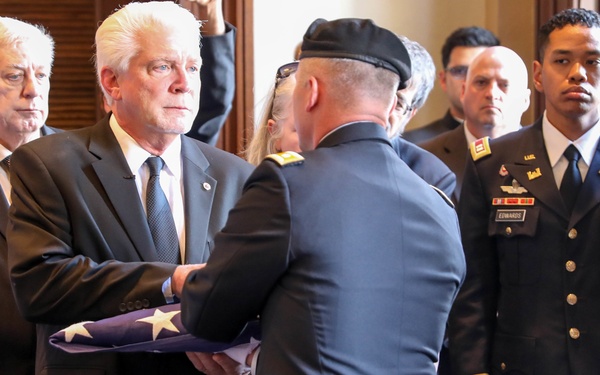 A military funeral ceremony is taking place indoors, likely in a government or official building. A uniformed military officer is presenting a folded American flag to a civilian man in a dark suit, while other military personnel and civilians stand solemnly in the background. A casket draped in what appears to be a flag or covering is partially visible in the foreground.
