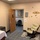 The image shows a small institutional room, likely a lactation/nursing room or a private consultation room in a medical or office facility. The room contains a cushioned rocking chair, a padded examination table or bench, and decorative wall elements including floral/sunflower decals. The open door reveals a hallway with a framed picture and a waiting area chair visible beyond.