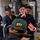 A group of military personnel, likely U.S. Navy sailors, are gathered in what appears to be a shipboard compartment or training space. One person in the foreground wears a green cap labeled 'ETT CG-57', suggesting this is aboard USS Lake Champlain (CG-57). A sailor is holding a red booklet or manual, and the group appears to be engaged in a briefing or training session.