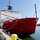 A large red and white vessel with hull number '20' is docked at a pier in calm waters. The ship features a reinforced red hull consistent with icebreaker design, yellow cranes, and extensive communications equipment on its superstructure. The vessel is moored with lines to yellow bollards on a concrete dock.