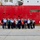 A group of approximately 20-25 people are posing for a group photograph in front of a large red U.S. Coast Guard vessel docked at a pier. The group includes individuals in civilian attire as well as at least two people in U.S. Coast Guard uniforms. The partial text 'U.S.' and 'GUA' (Guard) is visible on the red hull of the ship.
