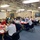 A formal meeting or briefing is taking place in what appears to be a shipboard conference room, with approximately 15-20 participants seated at red tables arranged in a U-shape or cluster formation. Several attendees are wearing face masks, suggesting this occurred during the COVID-19 pandemic period. The room features exposed ceiling infrastructure typical of a naval vessel, along with display screens and flags.
