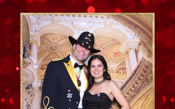 Two individuals pose for a photo booth picture at the St. Barbara's Day Ball 2020, held at Fort Hood, Texas. The man is wearing a U.S. Army dress uniform (Army Blues) with a black cavalry Stetson hat, gold aiguillette, medals, and rank insignia. The woman is wearing a black strapless formal gown with a choker necklace.