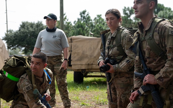 Military personnel in camouflage uniforms are conducting a field medical training exercise outdoors. A soldier numbered '07' is kneeling beside a medical training mannequin on a litter/stretcher, practicing casualty care procedures. Observers in both civilian-style PT gear and full combat kit with weapons and rucksacks are watching the exercise.