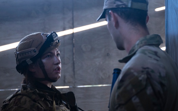Two military personnel are engaged in a conversation inside what appears to be a dimly lit indoor facility or training environment. The individual on the left is wearing full tactical gear including a combat helmet with mounted equipment, body armor, and multicam/OCP pattern uniform. The person on the right is wearing a patrol cap and the same pattern uniform without visible helmet.