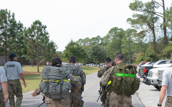 A group of military personnel in camouflage uniforms are walking along a paved road at what appears to be a military installation. Three individuals wearing gray shirts labeled 'CADRE' are accompanying several trainees or candidates carrying large rucksacks. The scene suggests a ruck march or physical training exercise as part of a military training program.