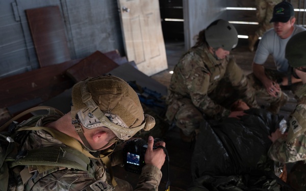 Military personnel in OCP (Operational Camouflage Pattern) uniforms are conducting what appears to be a tactical combat casualty care (TCCC) training exercise inside a wooden structure. One soldier in the foreground is using a camera or small device to document or review the scenario, while others attend to a simulated casualty on the floor. The scene includes multiple personnel with tactical gear, helmets, and medical supplies.