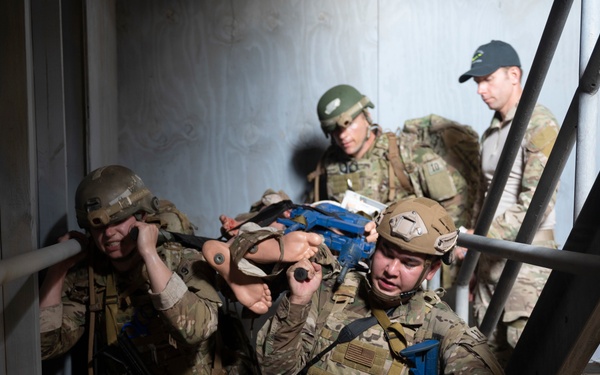 A group of military or law enforcement personnel in full tactical gear are conducting what appears to be a training exercise inside a stairwell of a building. The personnel are carrying a simulated casualty on a stretcher while navigating the confined staircase. The blue-colored training weapons and the presence of an observer/evaluator in civilian-style cap indicate this is a controlled training scenario rather than a live operation.
