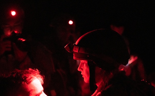 A nighttime scene illuminated by red light showing a helmeted individual in tactical gear facing and appearing to speak with or assist another person. The scene is dramatically lit with red-tinted light sources, creating high contrast against a very dark background. Multiple figures are visible in the background, some appearing to hold equipment.