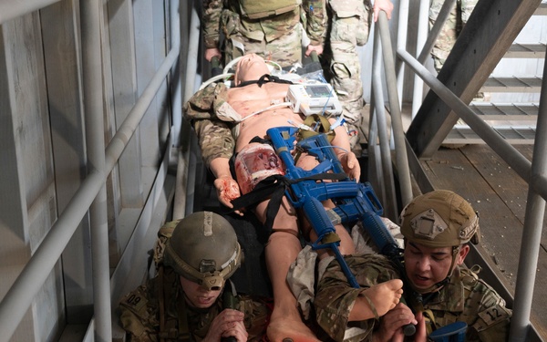 Military personnel in camouflage uniforms are conducting a casualty evacuation (CASEVAC) training exercise, carrying a simulated casualty on a litter down a stairwell. The 'casualty' is a mannequin or role-player with simulated wounds and a blue training rifle placed on top. This appears to be a realistic combat medical training scenario in a controlled environment.