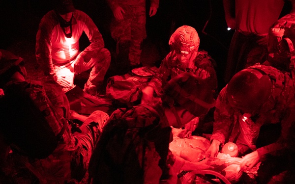 Military personnel are conducting a nighttime medical treatment or casualty care exercise under red light illumination. Multiple individuals in military uniforms and helmets are gathered around what appears to be a patient or casualty on the ground. The red lighting is consistent with tactical night operations to preserve night vision.