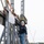 Two workers in hard hats are operating a large metal trebuchet or catapult machine at what appears to be a 'Punkin Chunkin' competition. A sign on the machine references 'Punkin Chunkin 2012 World Champion' and 'Western New York.' One worker in a black hoodie stands on the machine's stairway while another in an orange hoodie holds a green pumpkin or gourd at the base.