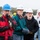 A group of workers or volunteers wearing hard hats and work gloves are gathered outdoors in a field or open area. They appear to be attending a briefing, demonstration, or safety meeting, standing attentively and listening. The setting suggests a construction, environmental, or community work event taking place in cool weather.
