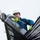 A worker named Bryan, identified by his jacket embroidered with 'Team Ethan' and 'The Phoenix', is working at height on a structure made of dark cables or rods. He is wearing safety equipment including a white hard hat, yellow high-visibility gloves, sunglasses, and a blue jacket. He appears to be handling a mechanical component with ropes/cables attached, likely performing rigging or installation work.