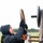 A man wearing a black North Face jacket with hood up and black gloves is working outdoors in cold, overcast conditions. He is lifting or positioning a large circular metal ring/weight plate onto a wooden and metal frame structure. A second person is partially visible in the lower right background.