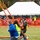 A group of young people wearing safety vests are operating what appears to be a large trebuchet or catapult device on a grassy field during an outdoor event. A pumpkin is visible suspended on the right side of the frame, suggesting this may be a pumpkin-launching competition or demonstration. The event appears to be organized, with orange safety fencing, a large white tent, and staff in high-visibility vests visible in the background.