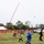 A group of participants are operating a large trebuchet or catapult-style launcher at what appears to be an outdoor fall festival or pumpkin-launching event. Several people wearing safety vests are pulling ropes to trigger the device, which has just launched a projectile (likely a pumpkin) visible in the upper right of the frame. Spectators watch from behind orange safety fencing in the background.