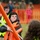 Two young people wearing safety vests are engaged in a physical team activity outdoors, appearing to pull on a rope near orange PVC pipe structures. The foreground subject wears an orange safety vest over a gray hoodie and is gripping a rope with visible effort. The event appears to be an organized outdoor competition or team-building exercise with spectators and orange fencing visible in the background.
