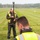 Two workers wearing high-visibility safety vests are conducting land surveying work in an open grassy field. The worker in the background is holding a surveying rod topped with a GPS/GNSS receiver unit, while a second worker in the foreground is out of focus. The setting appears to be a rural or semi-rural area with mowed grass and tree lines visible in the distance.