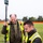 Two workers wearing high-visibility safety vests are conducting land surveying work in an open grassy field. The younger worker is operating a handheld data collector/controller device while the older worker holds a survey rod topped with a GNSS/GPS receiver antenna. Orange construction fencing and trees are visible in the background.
