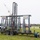A large trebuchet mounted on a trailer is captured mid-launch, with a projectile (likely a pumpkin) visible in the upper left arc of its trajectory. The machine bears NASAW and 'Punkin Chunkin' branding, indicating this is a competitive pumpkin-launching event. The trebuchet is set up on a grassy field at what appears to be an airfield or fairground, with spectators and vehicles visible in the background.