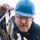 A middle-aged man wearing a blue hard hat and glasses is closely inspecting or working on a piece of industrial or mechanical equipment. He is dressed in a dark blue jacket and appears focused and concentrated on the task at hand. The background is overcast and bright white, suggesting an outdoor setting in cold or overcast conditions.