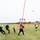 A group of people are operating a trebuchet or catapult-style launching device on an open grass field, having just launched a projectile (appearing to be a pumpkin or similar object) into the air. Several participants are pulling ropes to operate the mechanism while safety personnel in high-visibility vests oversee the activity. The setting appears to be an organized outdoor event, likely a pumpkin chunkin or harvest festival competition.