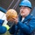 A man wearing a blue hard hat and blue jacket with 'ETHOS' and the name 'David' embroidered on it is carefully handling an orange pumpkin suspended by purple ropes or cords. The pumpkin has repeated markings printed on its surface, possibly 'E3'. The setting appears to be outdoors near a blue metal structure, suggesting a controlled drop or engineering experiment.