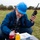 A middle-aged man wearing a blue hard hat and glasses sits on a grass field while holding a two-way radio/walkie-talkie up to his ear. He is looking down at a clipboard or document with a color-coded chart, with two beverage bottles placed nearby. In the background, a group of people in orange safety vests can be seen gathered along the perimeter of the field.
