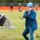 A middle-aged man wearing a blue hard hat, glasses, and a blue jacket is pulling a cable or wire attached to a piece of heavy equipment on a grassy field. He is wearing work gloves and appears to be participating in a groundbreaking ceremony or construction-related demonstration. Several observers and what appears to be a person in military uniform are visible in the blurred background.