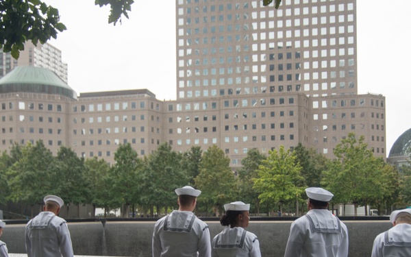 Six U.S. Navy sailors in white dress uniforms stand with their backs to the camera at the 9/11 Memorial in New York City, looking down at the engraved names on the memorial pool parapet. The scene appears to be taken on an overcast or rainy day, with the World Financial Center buildings and trees visible in the background. The image captures a solemn moment of remembrance at one of the two reflecting pools.