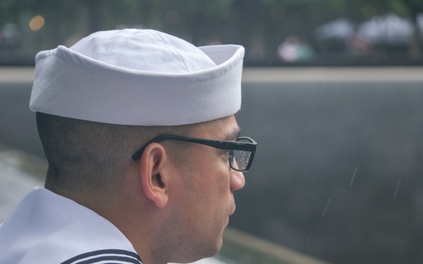 A U.S. Navy enlisted sailor in dress white uniform is photographed in profile, appearing to observe or pay respects at what appears to be the 9/11 Memorial in New York City. The sailor wears a command patch reading 'COMNAVREG MIDLANT' (Commander, Naval Region Mid-Atlantic) and a single star device on the shoulder. Rain is visibly falling in the background near the memorial's dark stone wall.