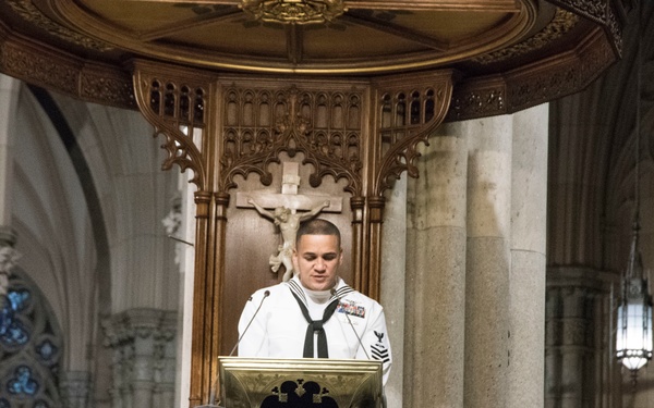 A U.S. Navy sailor in dress white uniform stands at an ornate white marble pulpit inside a large Gothic-style cathedral. He appears to be reading from a document or book placed on a brass lectern. The setting features elaborate stone carvings, arched columns, stained glass windows, and a wooden backdrop with a crucifix.