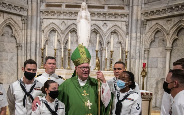 A Catholic bishop or cardinal in green and gold liturgical vestments poses with a group of U.S. Navy personnel in white dress uniforms inside an ornate Gothic-style cathedral or basilica. Several individuals are wearing black or blue face masks, suggesting this photo was taken during the COVID-19 pandemic period. The setting features marble walls, Gothic arches, gold candelabras, and a white statue in the background.