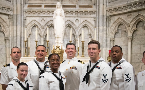 A group of approximately nine U.S. Navy personnel in white dress uniforms are posing for a formal group photograph inside what appears to be a large Catholic cathedral or basilica. They are standing in front of an ornate marble altar featuring a white statue, likely of the Virgin Mary, flanked by candles and Gothic architectural elements. The setting and attire suggest a ceremonial or commemorative occasion.