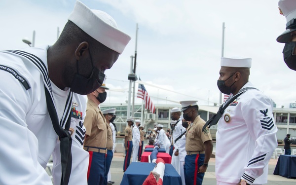 A military ceremony is taking place outdoors, likely at a naval installation or waterfront venue. Navy sailors in white dress uniforms and Marine Corps personnel in tan service uniforms are participating in what appears to be a formal presentation or award ceremony. Tables covered in red cloth are visible, and an American flag can be seen in the background.