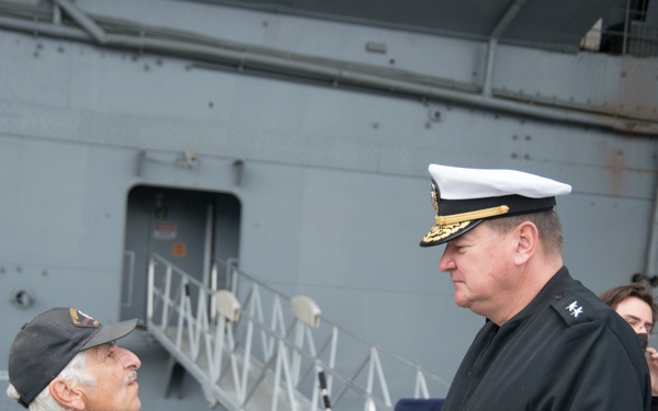 Two military personnel are engaged in conversation on what appears to be the deck or pier of a naval vessel. One individual wears a U.S. Army dress uniform with multiple patches and medals, while the other wears a senior U.S. Navy officer's uniform with a white-topped cap bearing gold insignia. A third person is partially visible in the background near the ship's gangway.