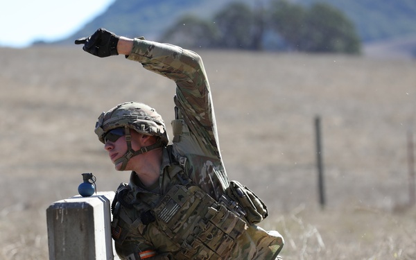 A U.S. Army soldier in full combat gear is crouched behind a concrete barrier in a dry, open field, appearing to simulate throwing a grenade during a training exercise. The soldier is wearing MultiCam (OCP) camouflage, a tactical vest, helmet, and sunglasses, with a practice grenade visible on top of the barrier. The setting appears to be a military training range based on the terrain and controlled environment.