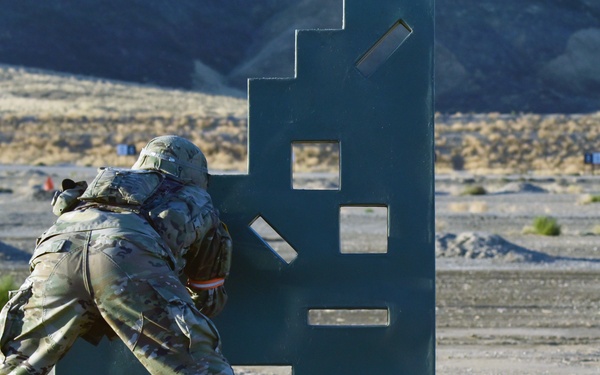 A soldier in full camouflage uniform and helmet is using a large metal barricade structure as cover while aiming a weapon at a military shooting range. The range features numbered target markers visible in the background across an arid, desert-like terrain. This appears to be a live-fire training exercise at an outdoor military qualification range.