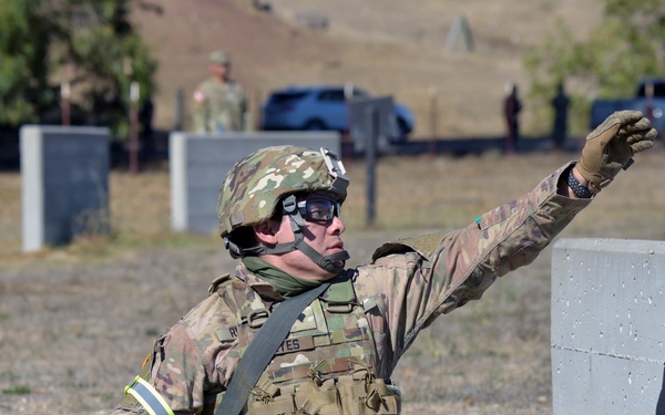 A U.S. Army soldier in full combat gear is seated on the ground beside a concrete block wall, in the act of throwing what appears to be a training grenade. The soldier is wearing OCP (Operational Camouflage Pattern) uniform, tactical vest, helmet, and protective eyewear. The setting appears to be a military training range with other personnel and vehicles visible in the background.