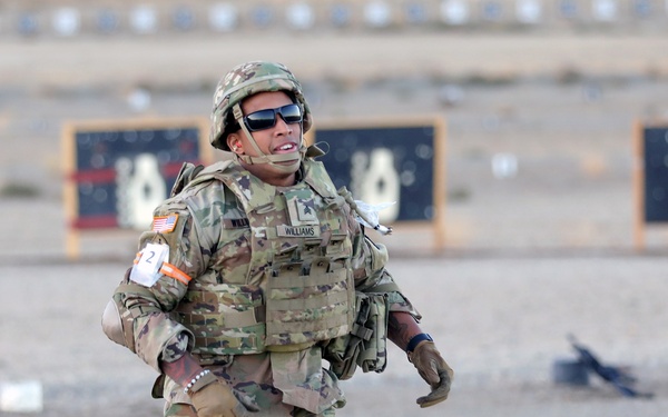 A U.S. Army soldier identified by name tape as 'Williams' is running across a gravel surface at what appears to be a military shooting range. The soldier is in full combat gear including a plate carrier, helmet, knee pads, and gloves, and is wearing a competition number '2' on their arm. Black silhouette shooting targets are visible in the background, suggesting this is a timed military competition or qualification event.