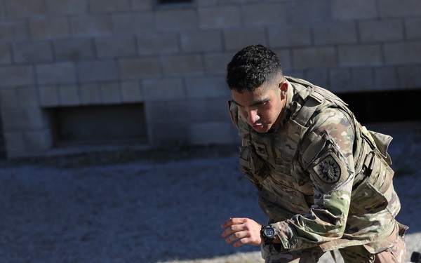 A soldier in OCP (Operational Camouflage Pattern) uniform is in a crouched, low-profile tactical position on dry, sparse ground near a cinder block structure. The soldier appears to be in motion or transitioning between positions during what appears to be a training exercise. A second person or equipment is partially visible on the ground in the background.