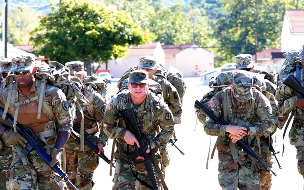 A large formation of military soldiers in full combat gear is conducting a ruck march on a paved road within what appears to be a military installation. The soldiers are wearing Operational Camouflage Pattern (OCP) uniforms, carrying rucksacks and M4-style rifles. The setting includes military-style buildings, trees, and a visible stop sign in the background.