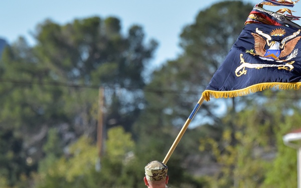 A U.S. Army soldier in MultiCam/OCP camouflage uniform is carrying a dark blue military unit colors (guidon/organizational flag) with gold fringe and an eagle emblem, walking along a road on what appears to be a military installation. The flag is extended outward on a wooden staff and appears to be in motion. The setting includes trees, mountains in the background, military-style buildings, and a utility truck.