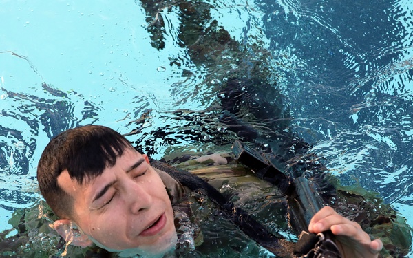 A young male soldier in wet camouflage uniform is swimming in what appears to be a pool while holding a rifle above the water. His eyes are closed and mouth slightly open, suggesting physical exertion. This appears to be a military water survival or combat swimming training exercise.