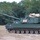 The image shows a self-propelled howitzer, consistent with the M109 Paladin series, painted in olive drab green and positioned on an open sandy field. The vehicle is stationary with its long 155mm cannon barrel extended forward-left. The setting appears to be a military training area or firing range, likely in the southeastern United States based on the vegetation.